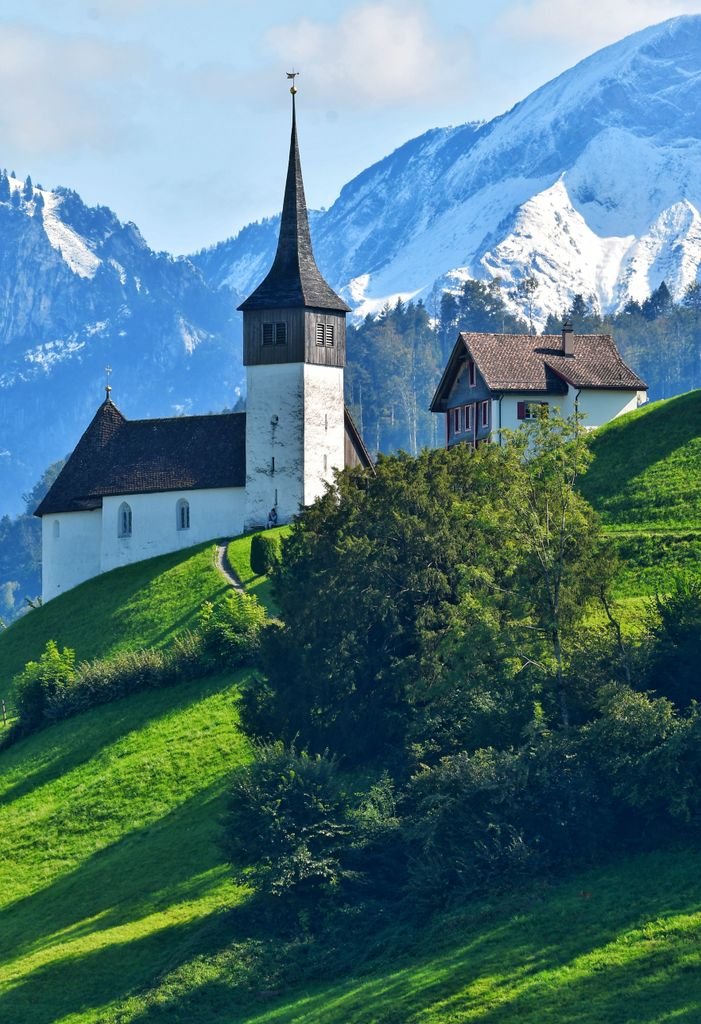 Picturesque Alpine church on lush green hills with snowy mountains in the background.